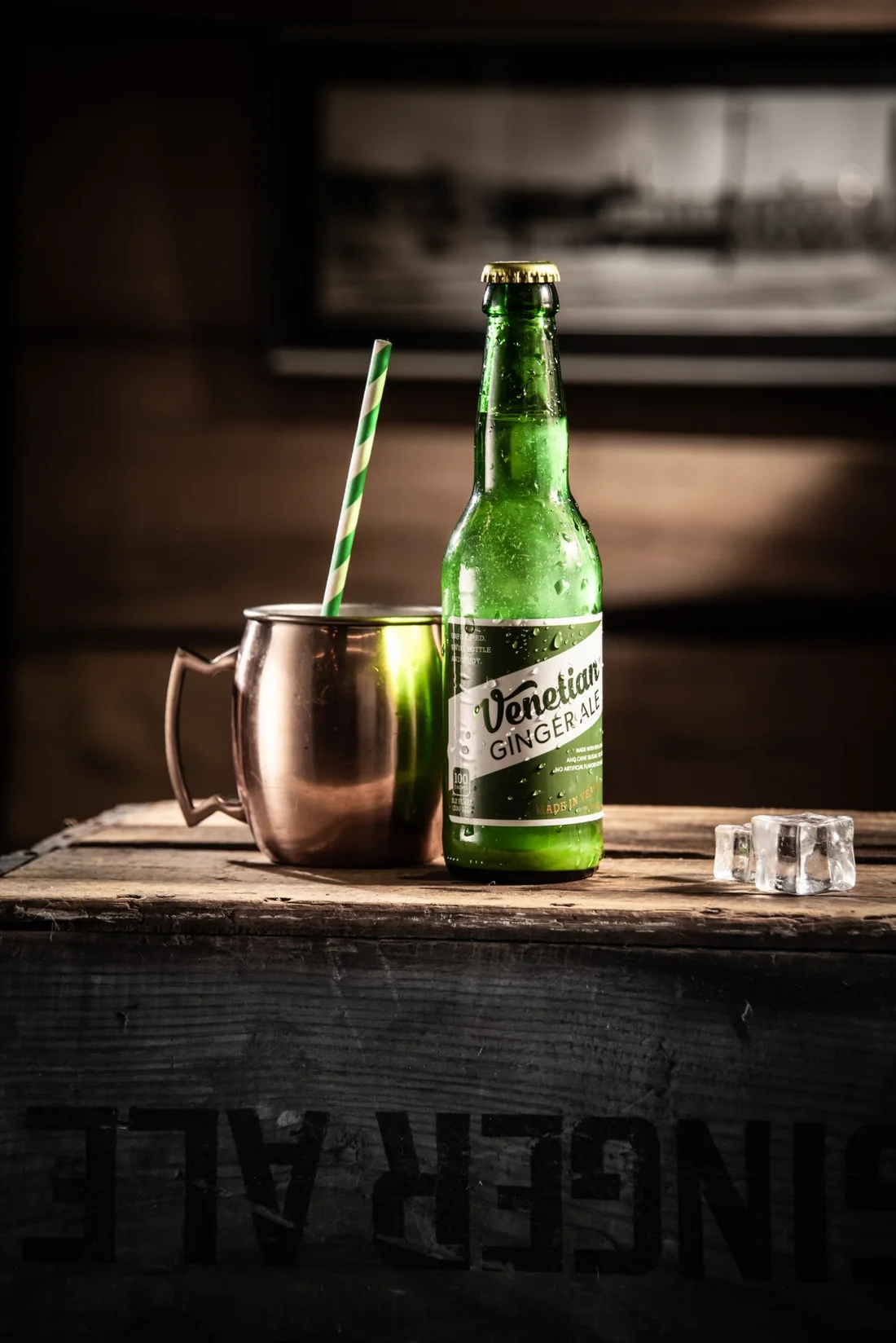 A chilled bottle of Venetian Ginger Ale beside a copper mug and striped straw, lit low against a dark wooden crate.