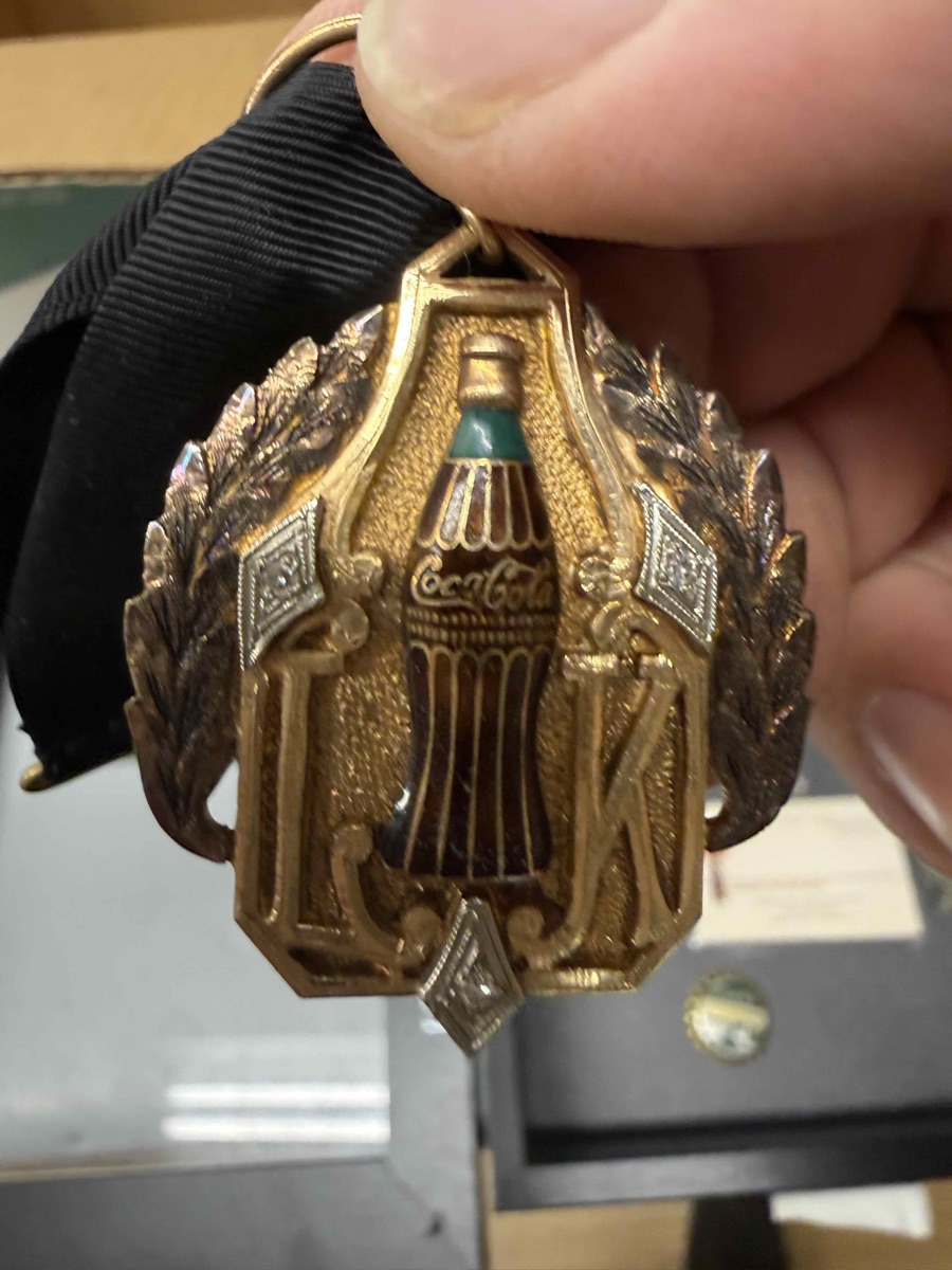 Gold Coca-Cola 100,000 Gallon Club medallion set with small diamonds, an enamelled Coke bottle at its centre, hanging from a black ribbon