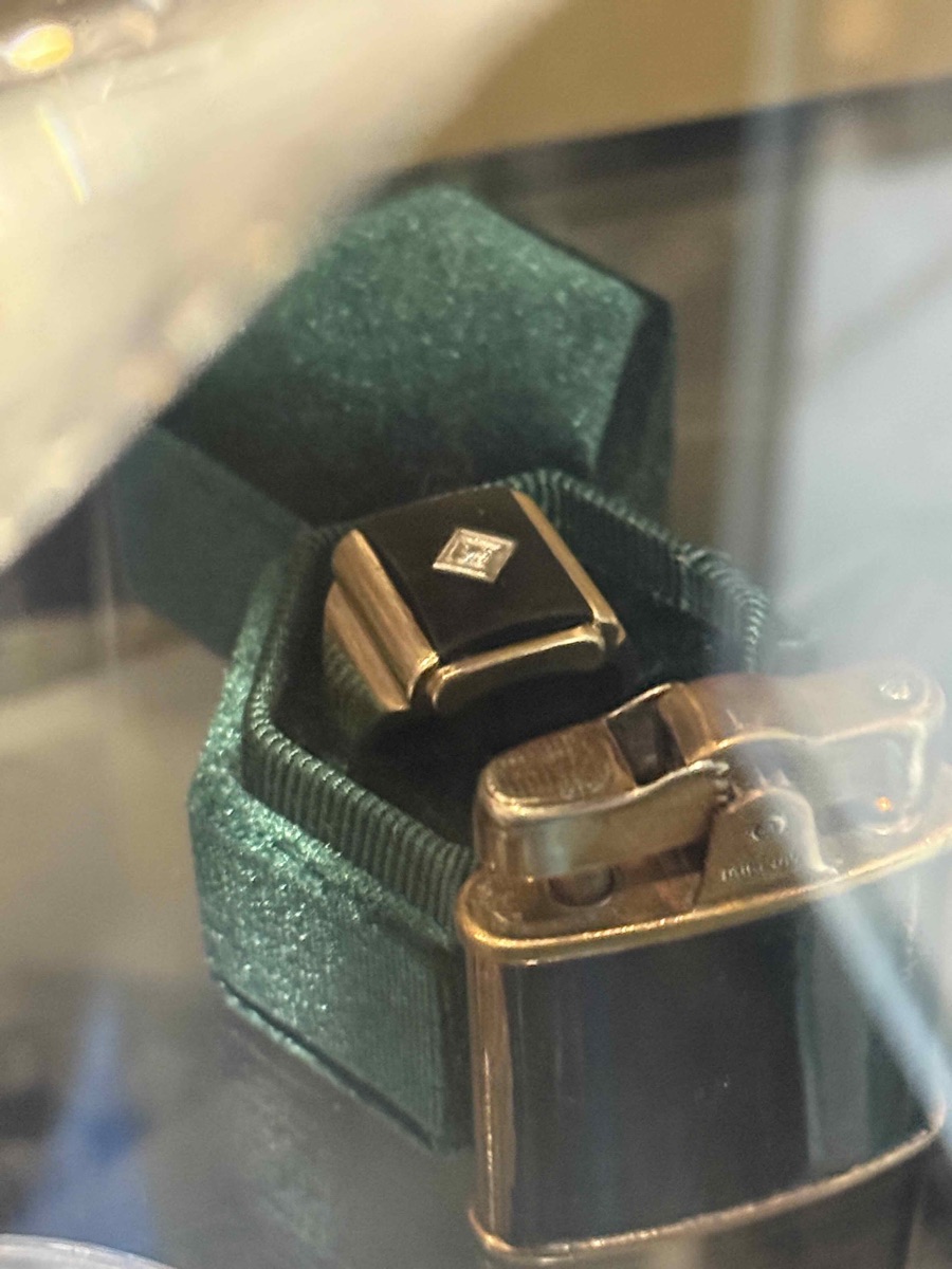 Gold ring with a black onyx face and a single diamond, sitting in a green velvet box beside a vintage lighter