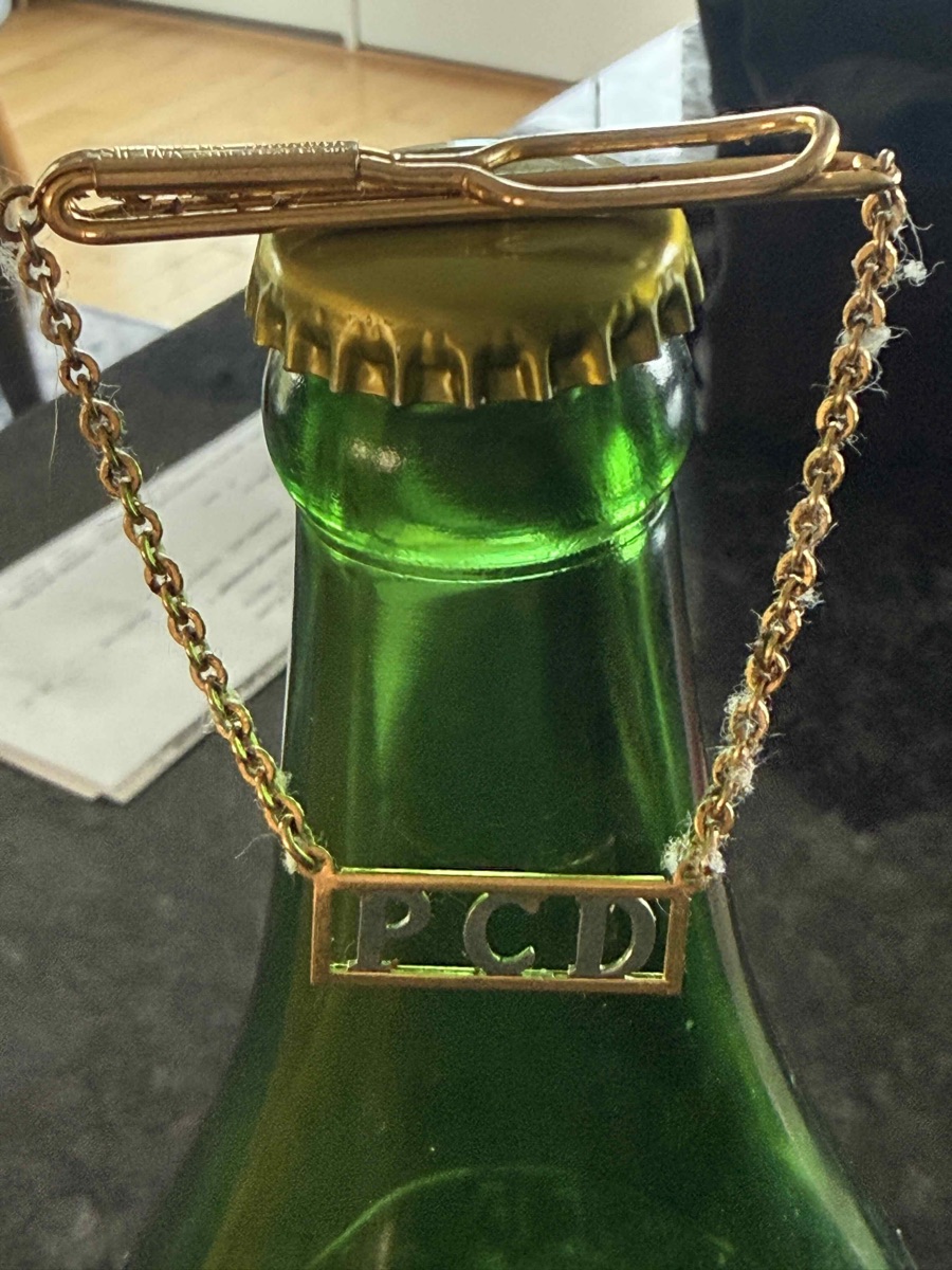 Gold tie clip with a chain and a bar monogrammed P.C.D., resting across the neck of a green Venetian bottle