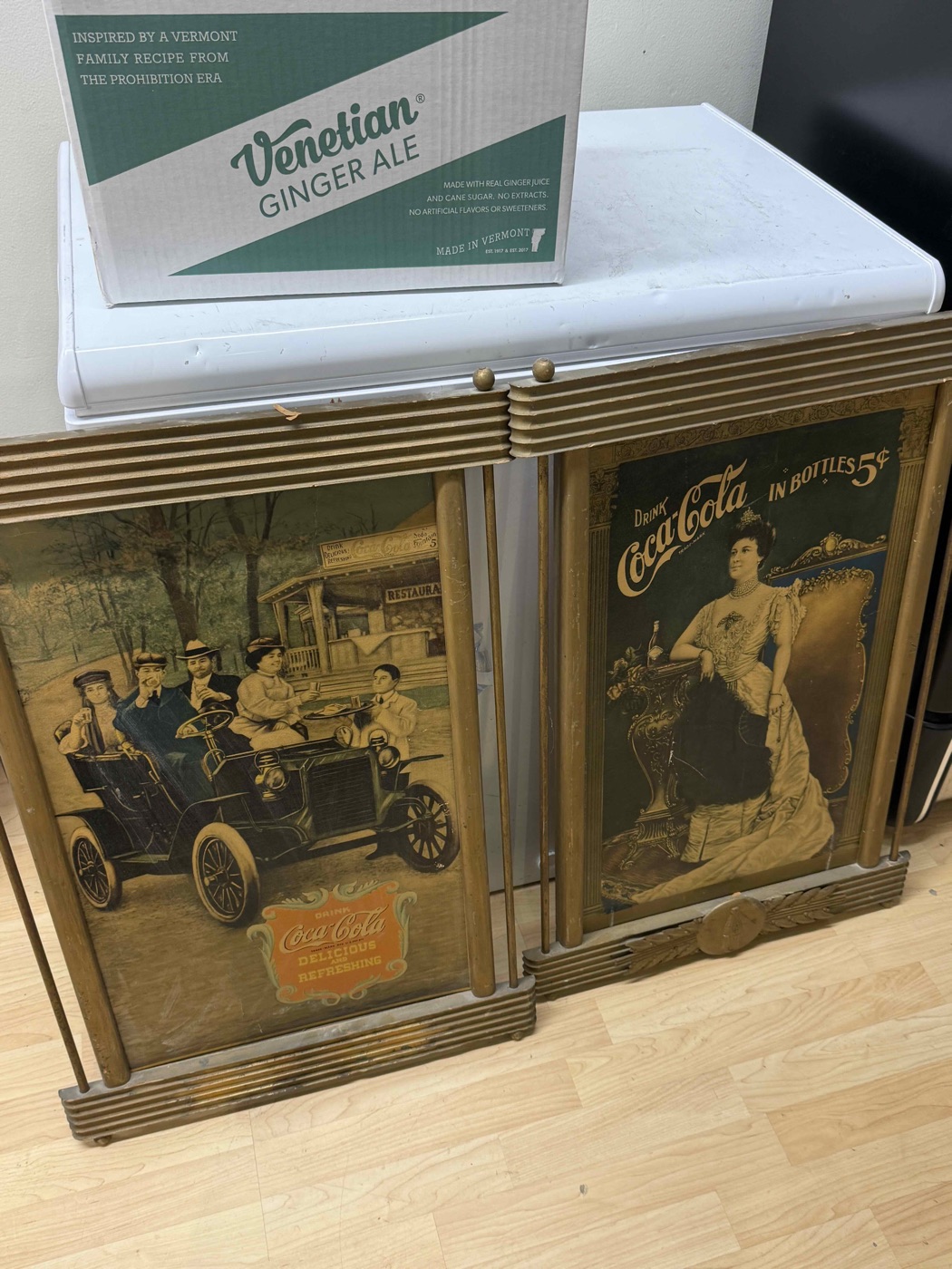 A modern Venetian Ginger Ale shipping case resting on top of an antique Coca-Cola display cabinet with illustrated glass panels