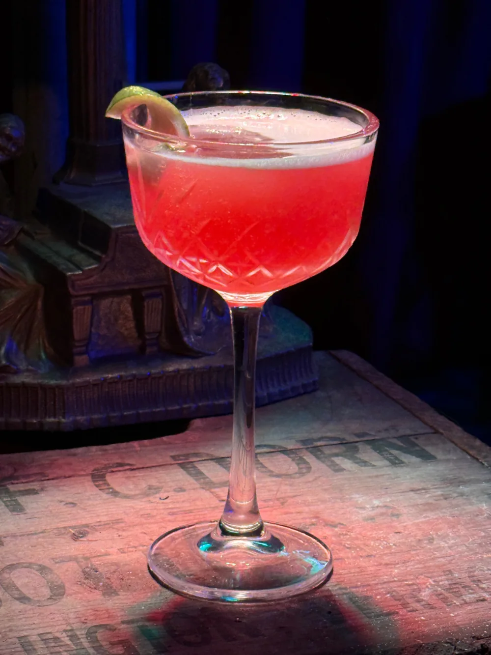 A red cocktail served up in a cut-crystal coupe with a lime wheel, resting on a wooden F.C. Dorn shipping crate.