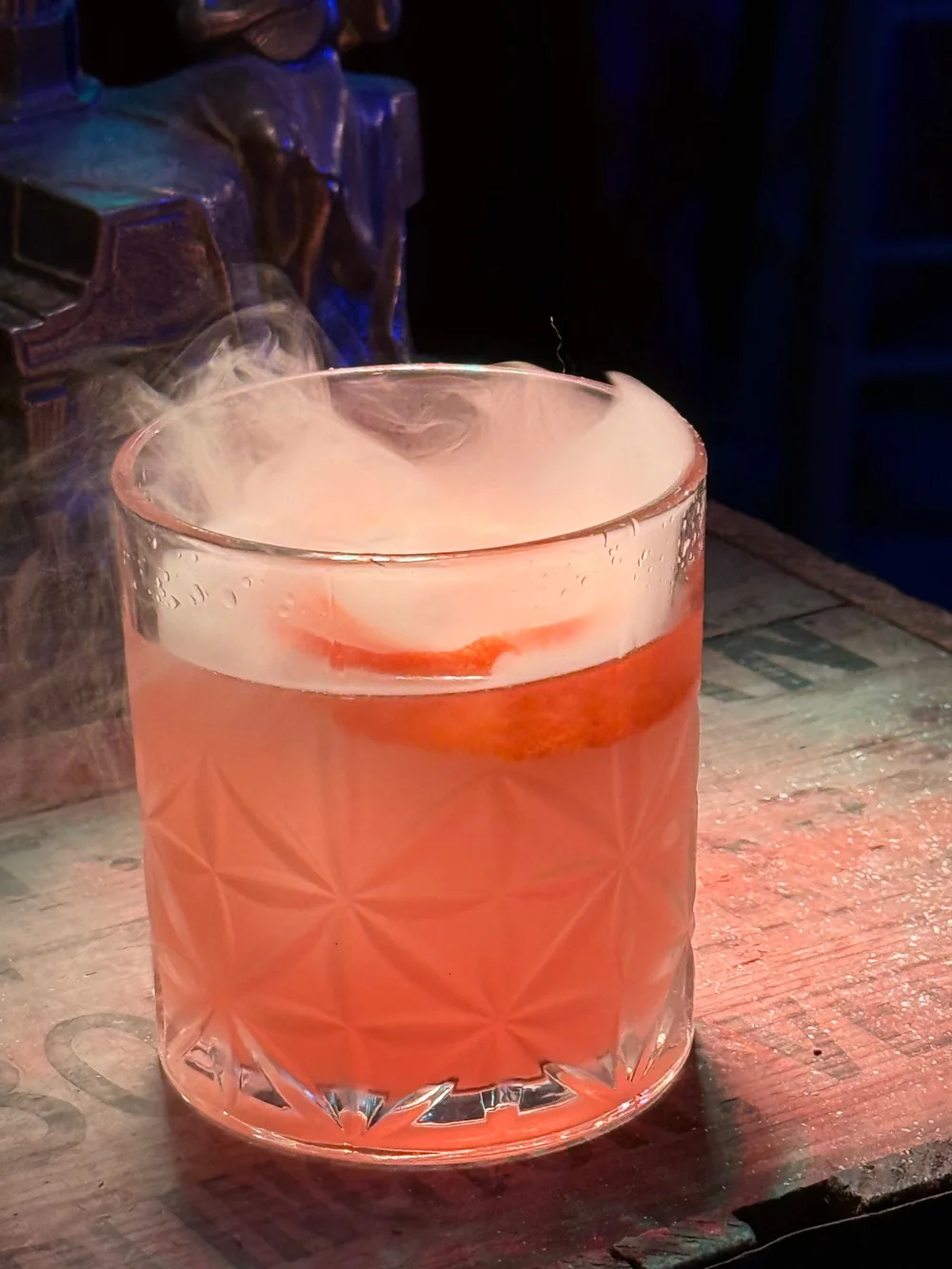 A grapefruit cocktail in a faceted rocks glass, smoke settling over the surface, on the same Dorn crate.