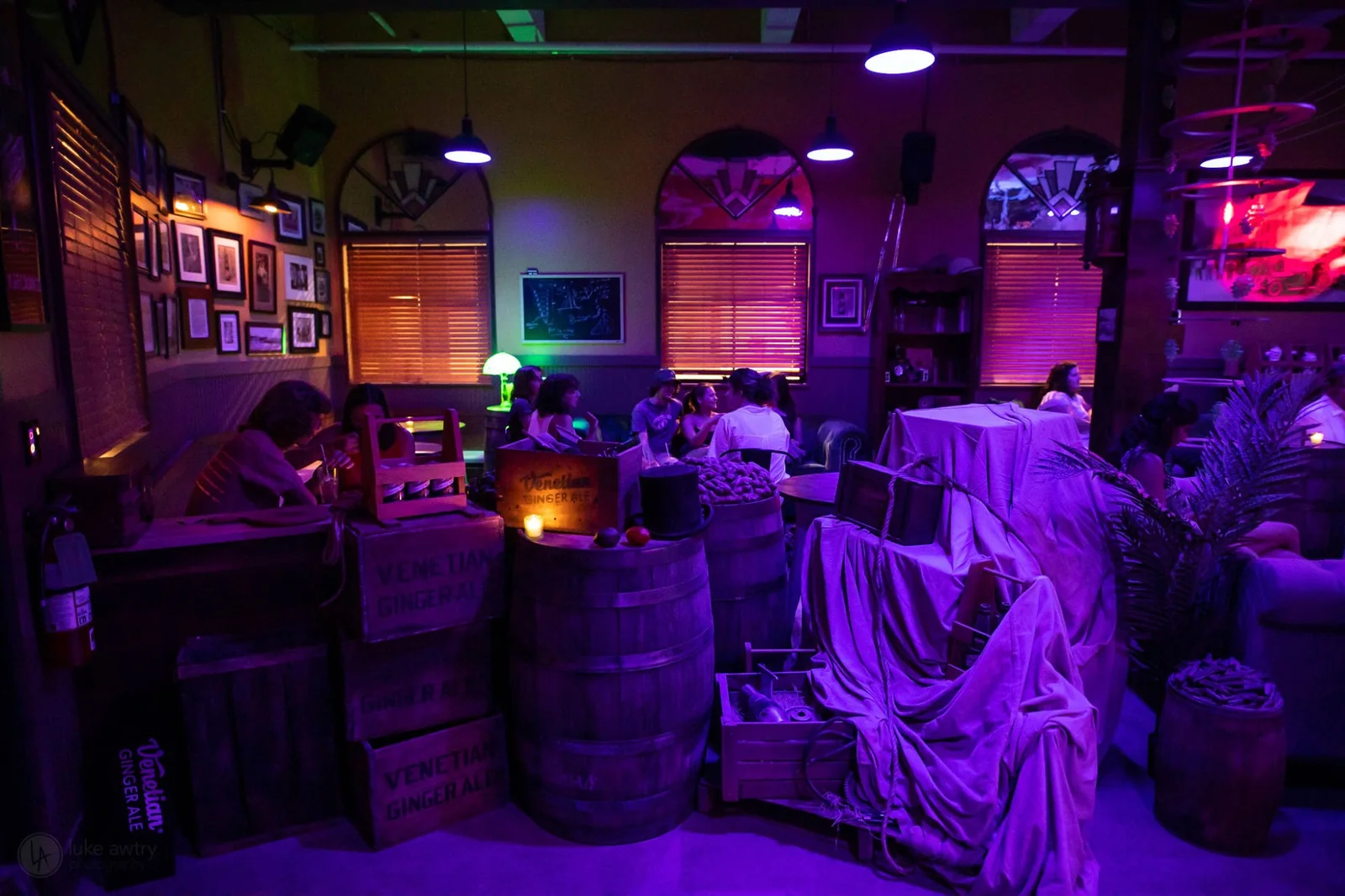 The Venetian Soda Lounge interior under purple and green light, with stacked Venetian Ginger Ale crates and barrels in the foreground and guests seated at tables beyond.