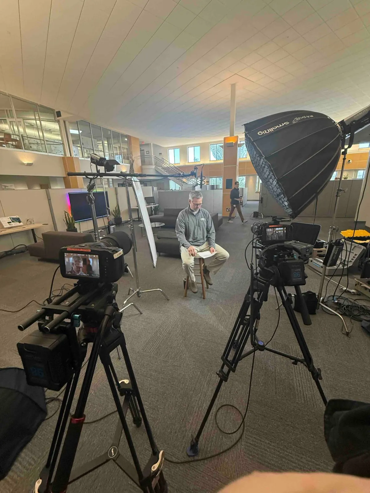 A subject seated on a stool in a corporate atrium, lit with a large softbox, two cameras running.