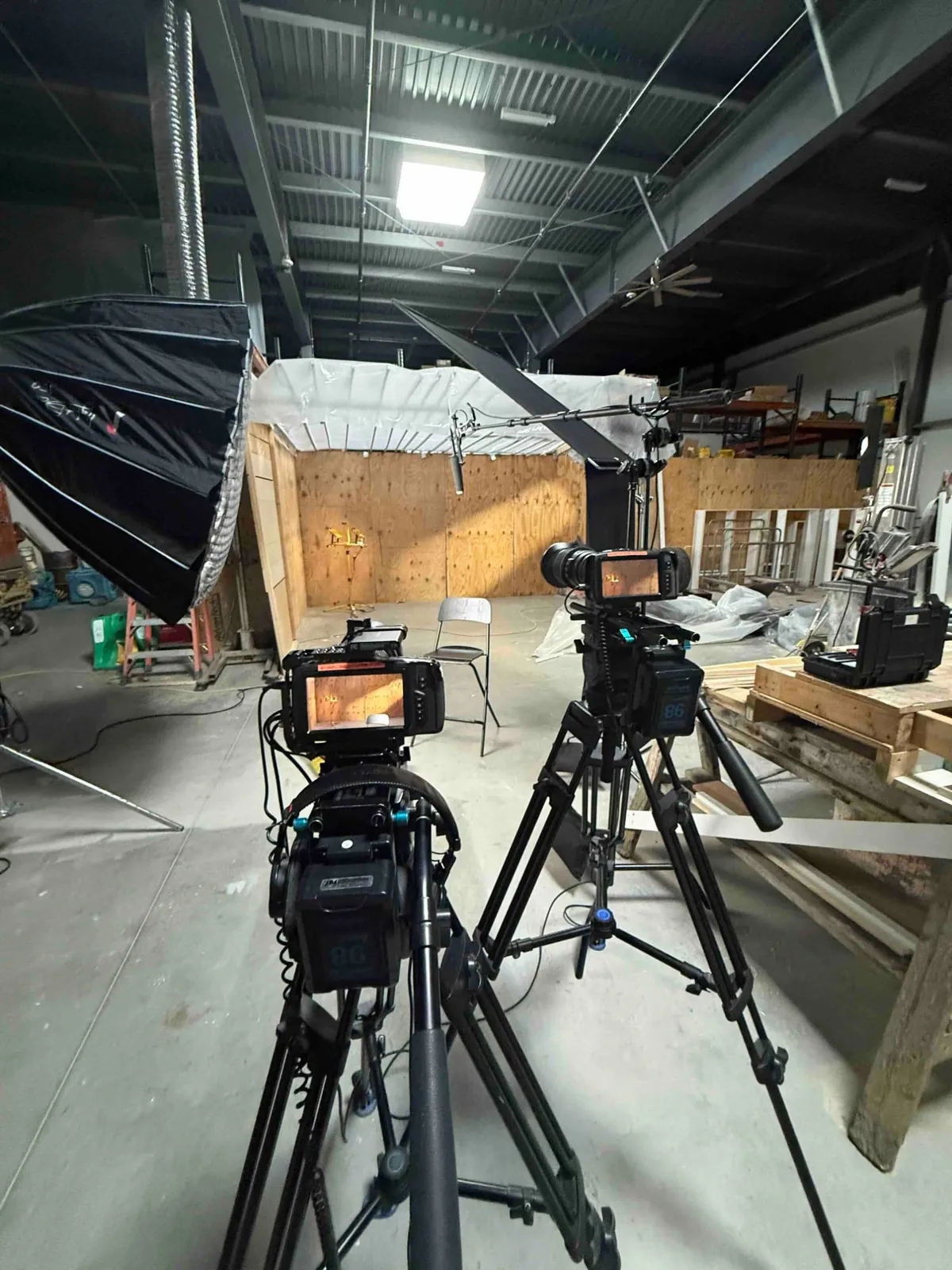 Two cameras and lighting set up in a warehouse in front of a plywood set build.