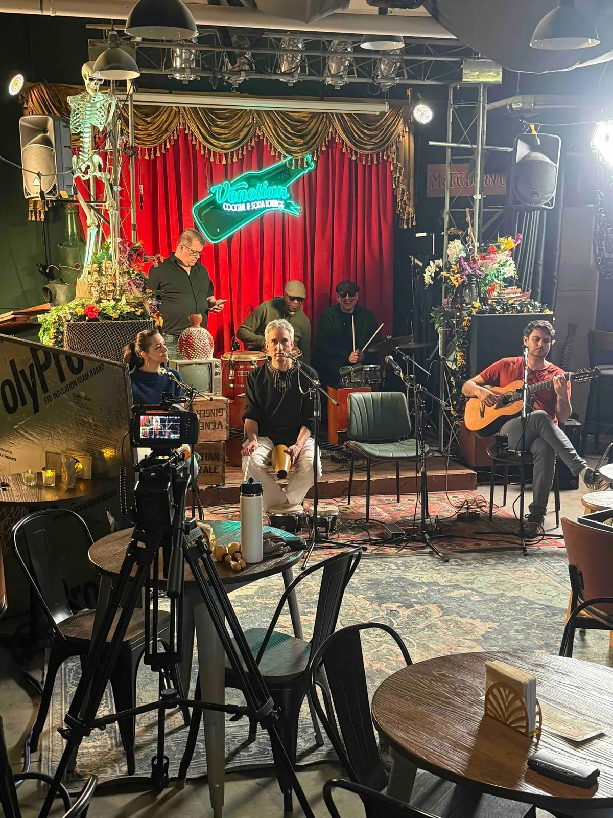 A band performing on the Venetian Soda Lounge stage under the neon bottle sign, camera on sticks in the foreground.