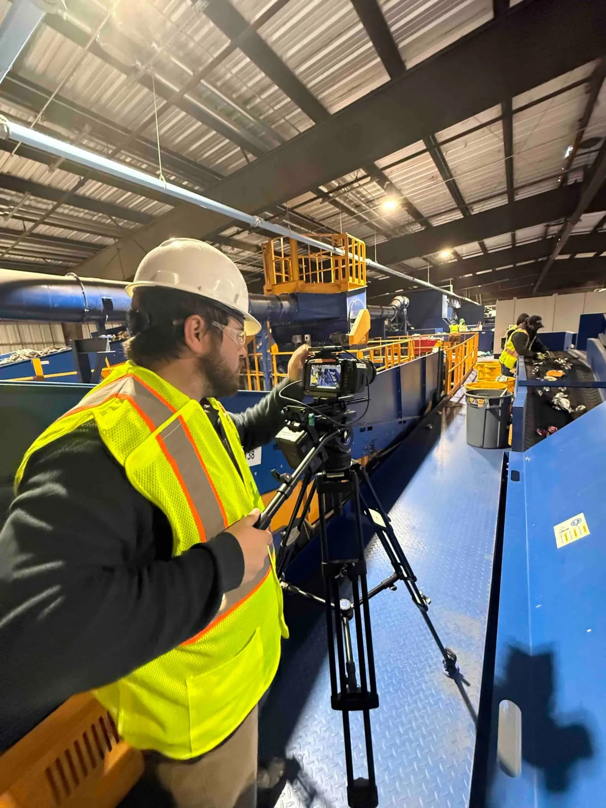 A camera operator in a hard hat and hi-vis vest filming a materials recovery facility sorting line.