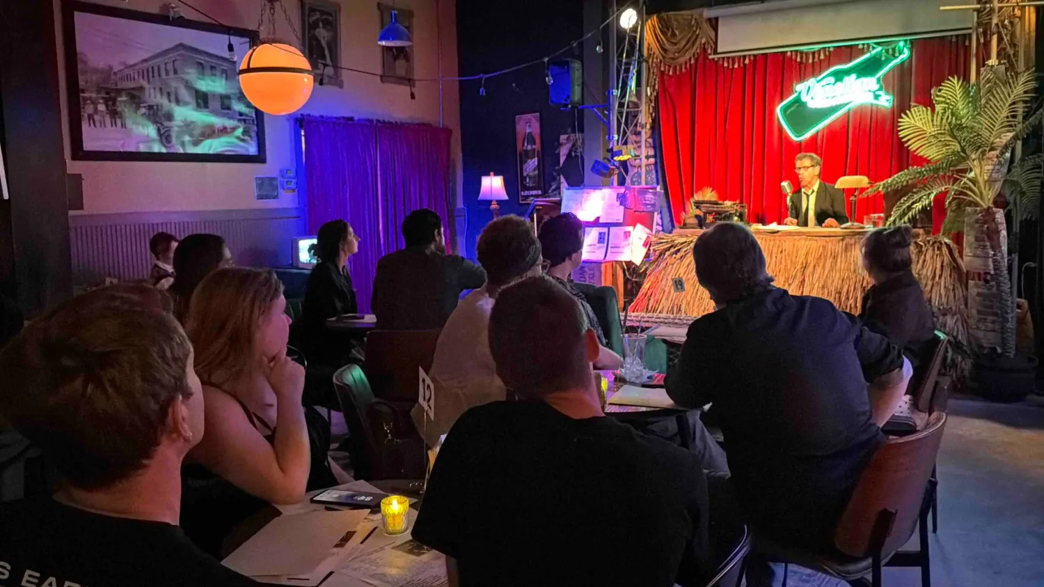 The performer seated at a desk on the lounge stage under a neon Venetian Ginger Ale sign, with the audience seated at tables watching.
