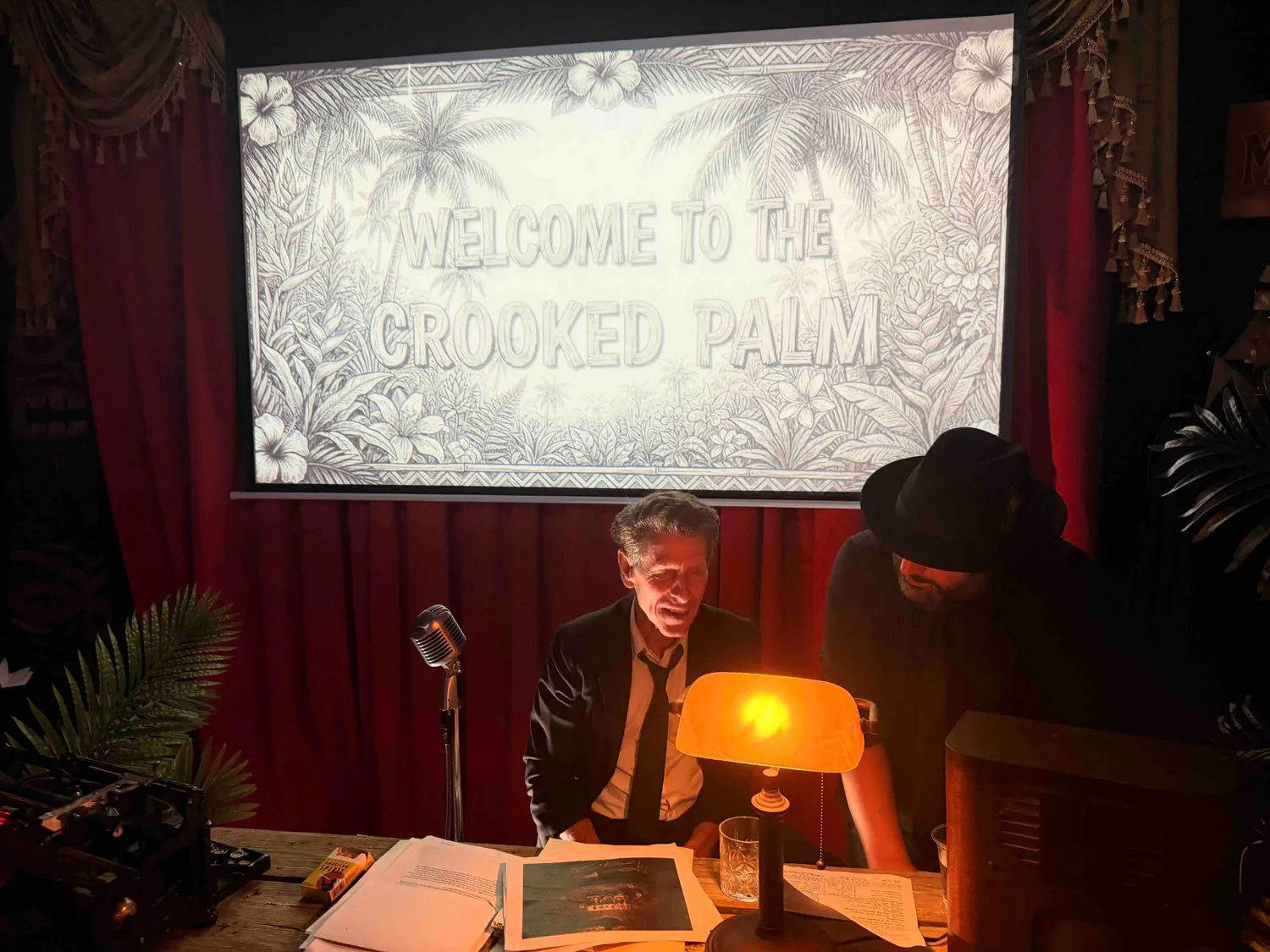 An actor in a dark suit and tie seated at a desk with a vintage microphone, banker's lamp, typewriter and scattered evidence papers, beneath a projected sign reading Welcome to the Crooked Palm.