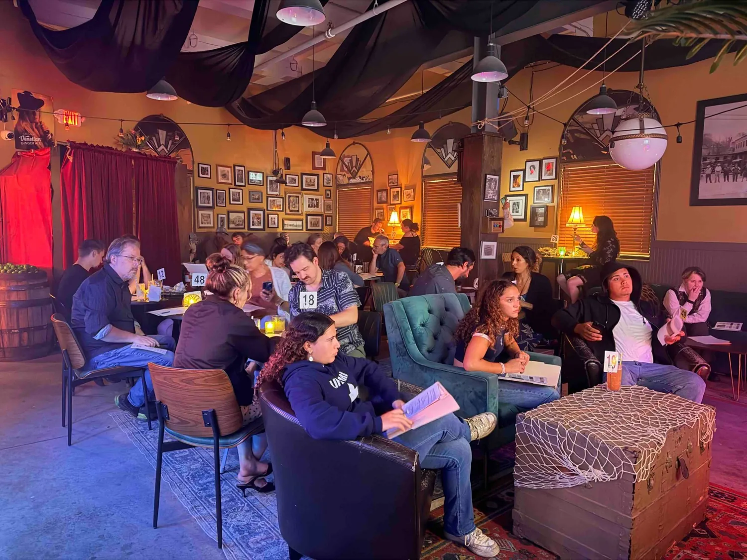 A wide view of the lounge filled with guests at numbered tables, drinks and printed case files in front of them.